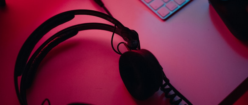 A dimly lit gaming setup with a monitor displaying a game's menu on the screen. A mechanical keyboard sits in front of the monitor. A pair of over-ear headphones and a game controller rest on the desk. The scene is bathed in a red and blue light, creating a moody gaming atmosphere.