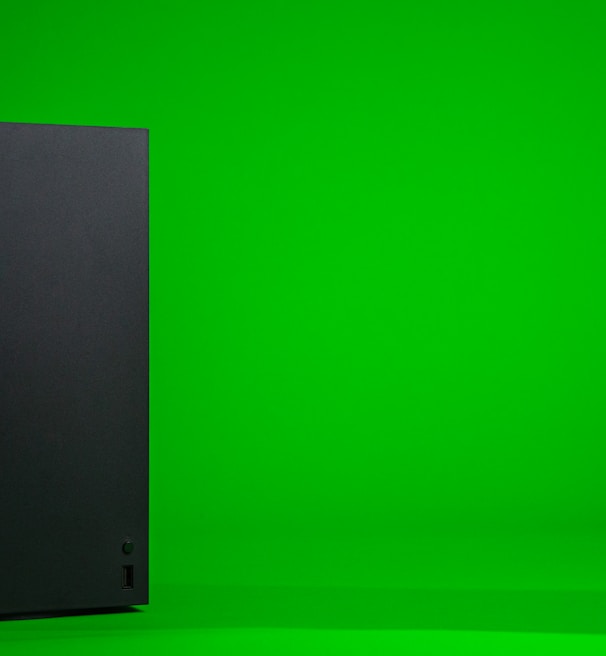 A black, rectangular gaming console stands upright against a vibrant green background. The console has a simple, sleek design with minimalistic features including a disc slot and a few buttons.