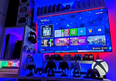A modern gaming setup featuring a large wall-mounted screen displaying a gaming console interface. The room is decorated with gaming-themed items such as controllers and collectibles. The ambient lighting includes shades of blue and red, creating a vibrant atmosphere.
