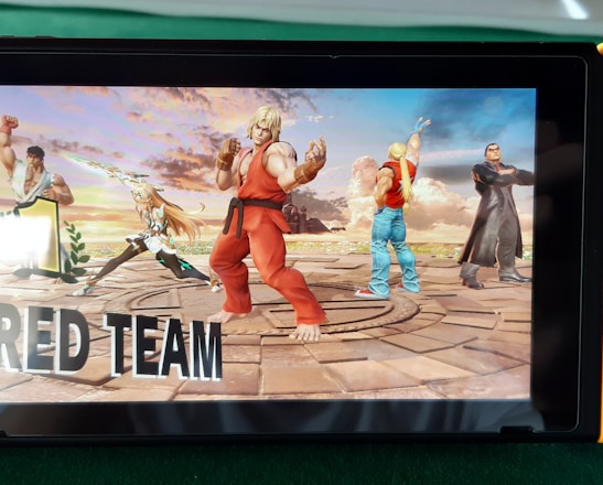 A handheld gaming console displays a screenshot of a fighting game featuring cartoonish characters in dynamic poses. The characters are standing in a virtual arena setting with a sky and cloud backdrop. The console is placed on a green surface.