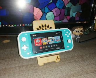 A turquoise handheld gaming console is placed on a wooden stand in front of a computer monitor displaying geometric shapes. To the right, there is a small figurine of a person wearing glasses and a suit. The gaming console screen shows a menu with various game icons.