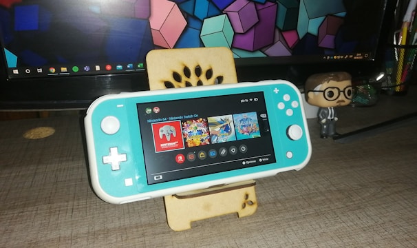 A turquoise handheld gaming console is placed on a wooden stand in front of a computer monitor displaying geometric shapes. To the right, there is a small figurine of a person wearing glasses and a suit. The gaming console screen shows a menu with various game icons.