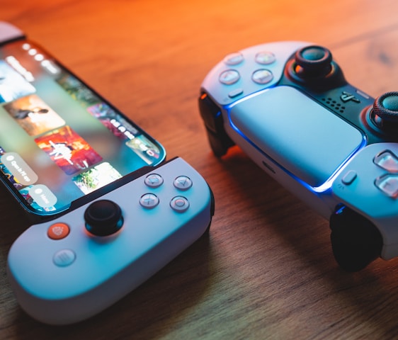 A gaming scenario featuring a smartphone with a game interface next to two controllers on a wooden surface. The smartphone displays vibrant images and icons, while the controllers feature a sleek, modern design with buttons and joysticks prominently visible. Warm lighting enhances the scene.