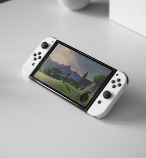 A portable gaming console with a screen showing a video game landscape, resting on a light-colored surface. The console has white controllers attached to either side of the screen. Nearby, there seems to be a white docking station or accessory.