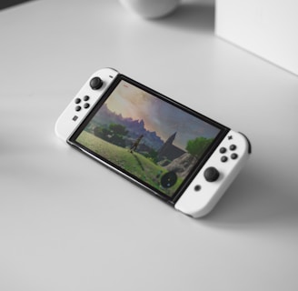 A portable gaming console with a screen showing a video game landscape, resting on a light-colored surface. The console has white controllers attached to either side of the screen. Nearby, there seems to be a white docking station or accessory.