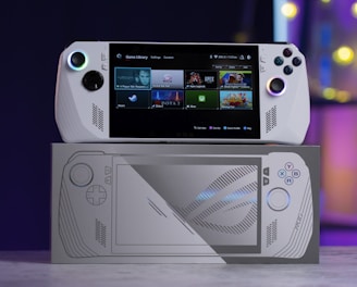 A handheld gaming console with a screen displaying a game library menu is placed on top of its packaging. The console features various buttons, a joystick, and a circular light around the left joystick. The background is blurred with purple, yellow, and blue tones.
