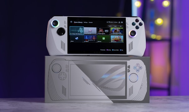 A handheld gaming console with a screen displaying a game library menu is placed on top of its packaging. The console features various buttons, a joystick, and a circular light around the left joystick. The background is blurred with purple, yellow, and blue tones.