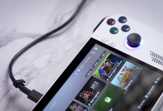 A handheld gaming console with a white body and colorful buttons features a screen displaying various game icons. The device is connected to a power source via a black cable and rests on a marble surface. The interface shows games like Apex and Street Fighter.