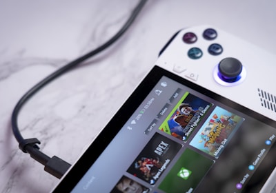 A handheld gaming console with a white body and colorful buttons features a screen displaying various game icons. The device is connected to a power source via a black cable and rests on a marble surface. The interface shows games like Apex and Street Fighter.