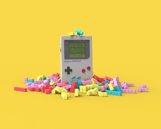 A vintage handheld gaming console surrounded by colorful, block-like pieces reminiscent of a classic puzzle game. The console has a small screen displaying a game in progress, with two red buttons and a directional pad. The vibrant pieces are scattered around it, creating a playful and nostalgic atmosphere.