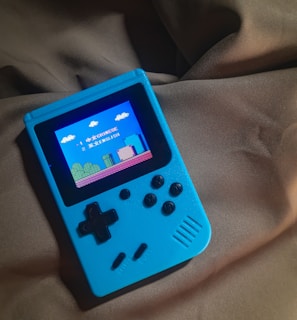 A handheld gaming device with a bright blue casing rests on a soft, dark fabric background. The screen displays a pixelated video game scene with options to select either Chinese or English language. The layout includes a classic D-pad and buttons, reminiscent of retro gaming consoles.